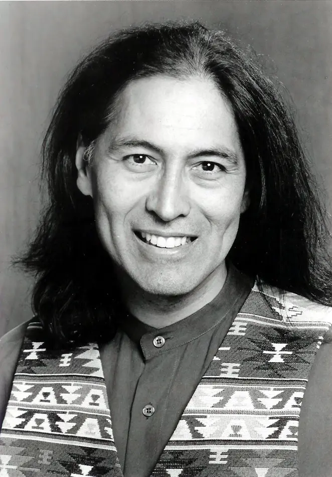 A black and white headshot of Val with long hair, clean shaven, and wearing a patterned vest. A wide smile sits on his face.